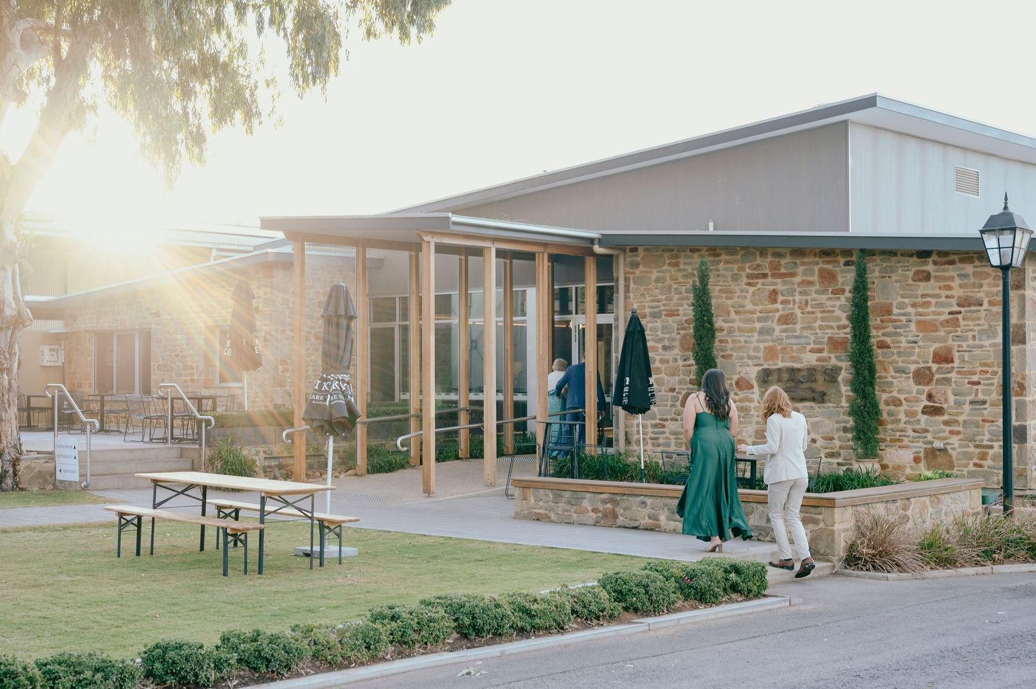 Modern stone building exterior with patio seating and green umbrellas during golden hour sunset.