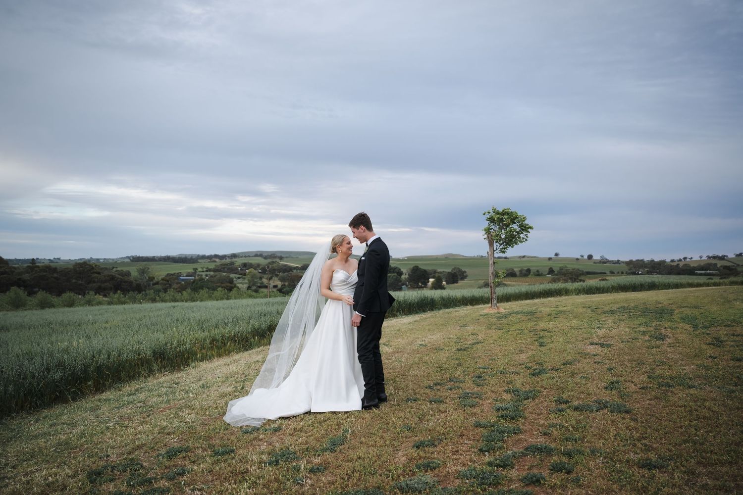 A couple embraces in an open field with rolling hills and cloudy skies in the background.