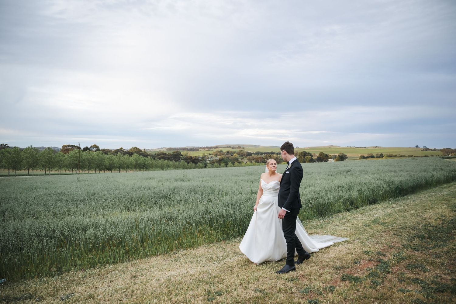 Couple walking together through scenic wheat fields on a sunny day with rolling hills in the background.