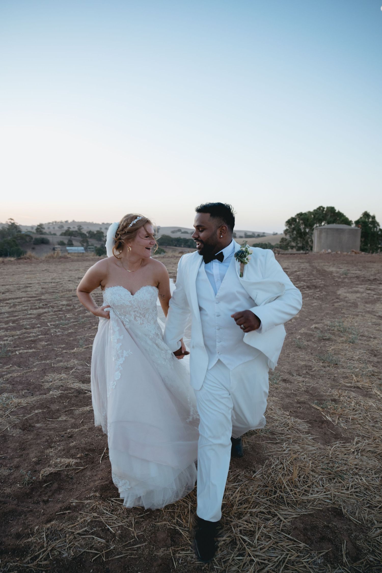 A romantic wedding couple poses together in a field at sunset wearing white formal attire.