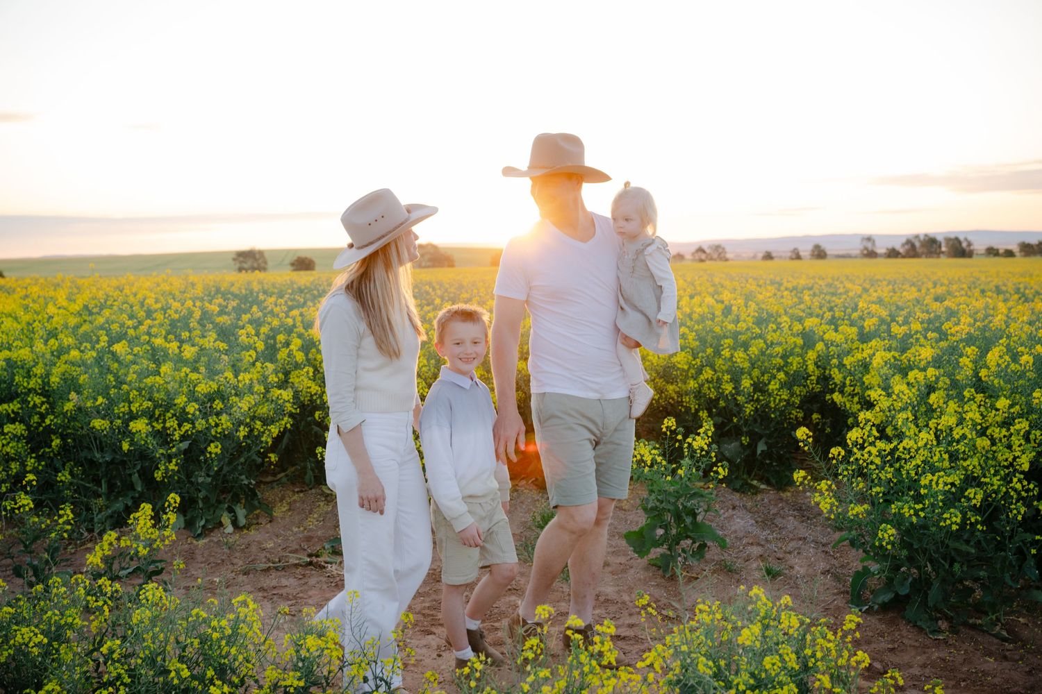 Family in white clothing walks through vibrant yellow canola fields during golden sunset hour.