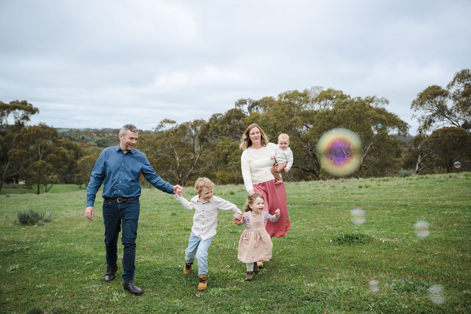 A family runs and plays together in a green field with eucalyptus trees in the background on an overcast day.