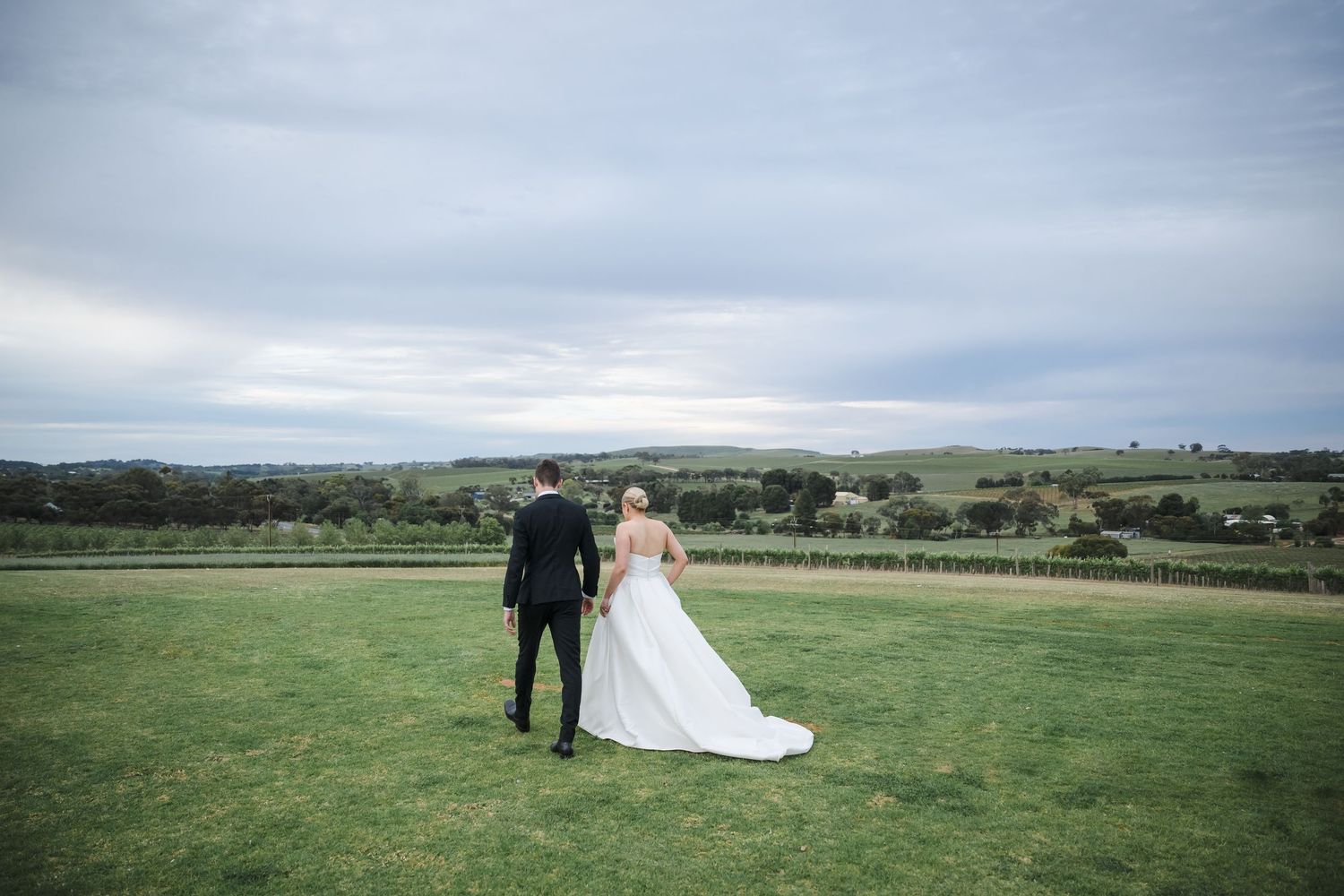 Wedding couple walks together across a scenic green meadow under cloudy skies.