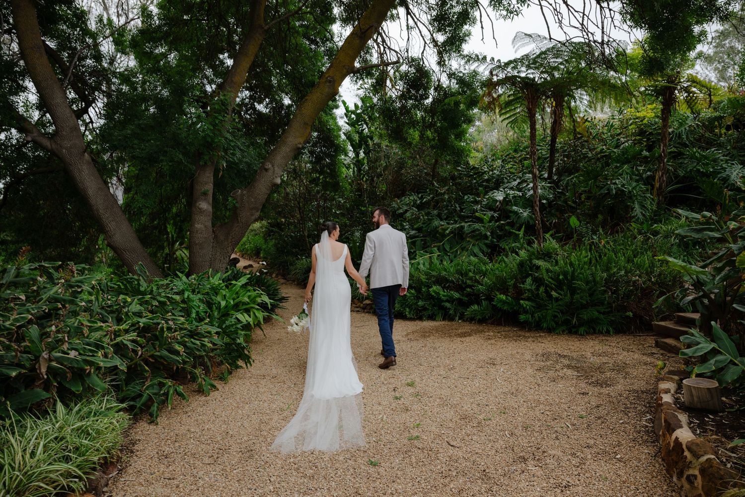 A couple walks down a rustic dirt path surrounded by lush green trees and foliage in a garden setting.