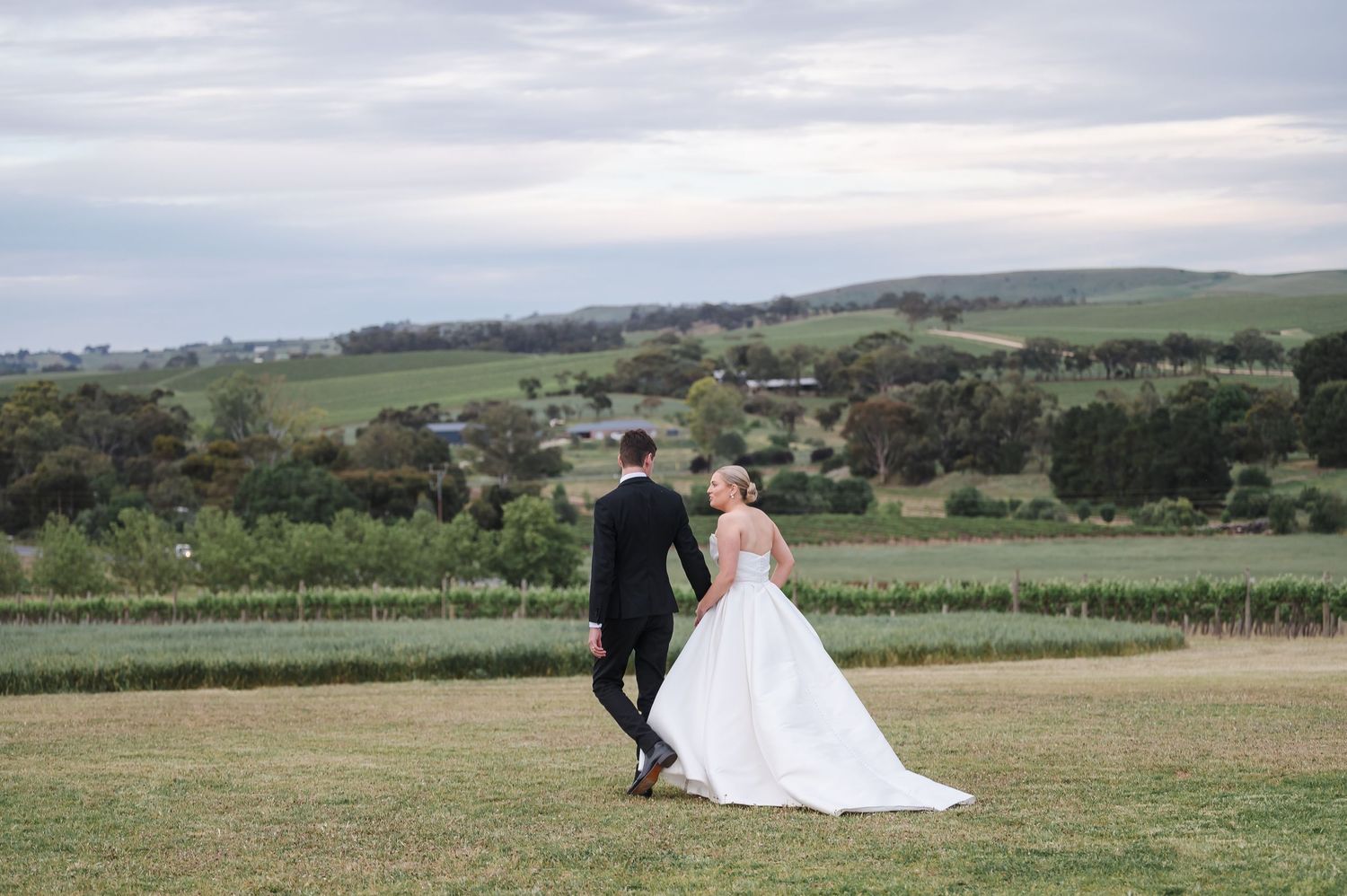 Newlywed couple sits together on grassy hilltop overlooking scenic countryside landscape at sunset.