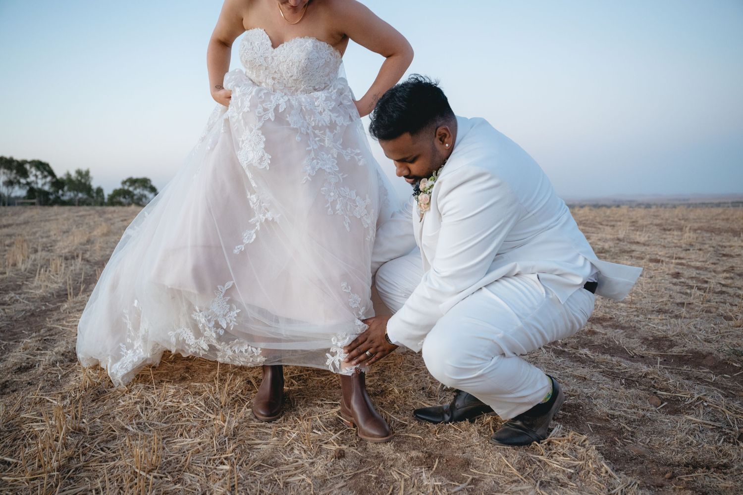 Couple in wedding attire crouches together on a sandy beach at sunset creating romantic moments.