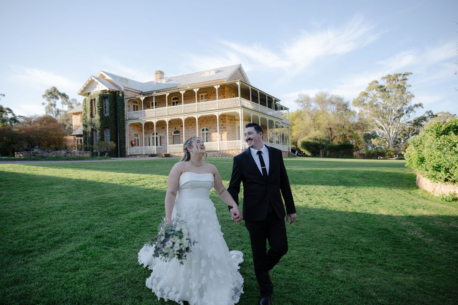 A wedding couple poses on a lush green lawn in front of a historic Victorian-style mansion with wraparound porch.