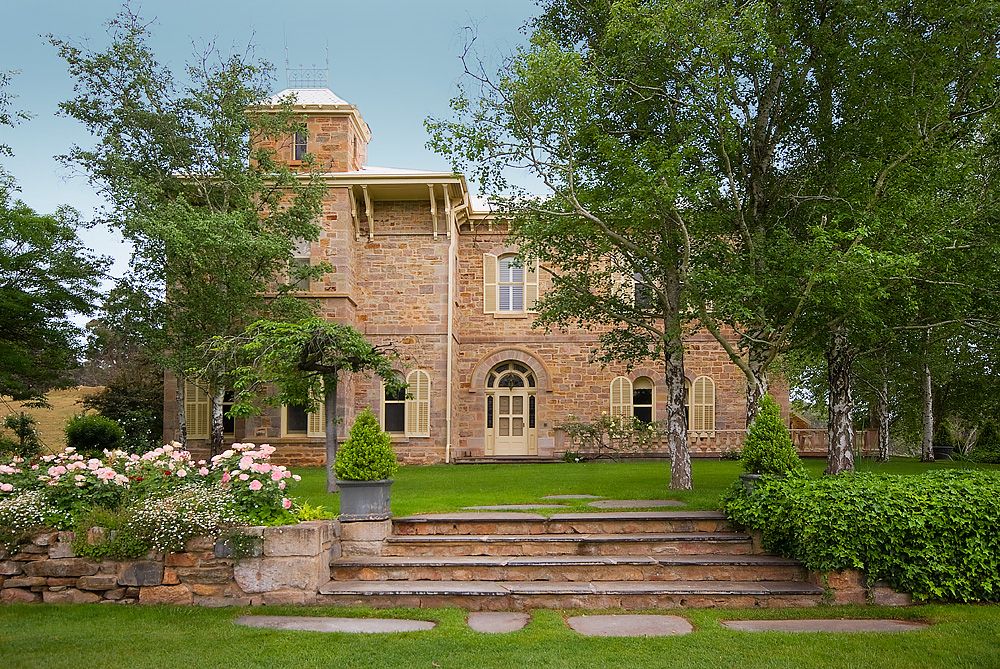 Elegant stone mansion with arched doorway and landscaped garden features stone steps and mature trees.