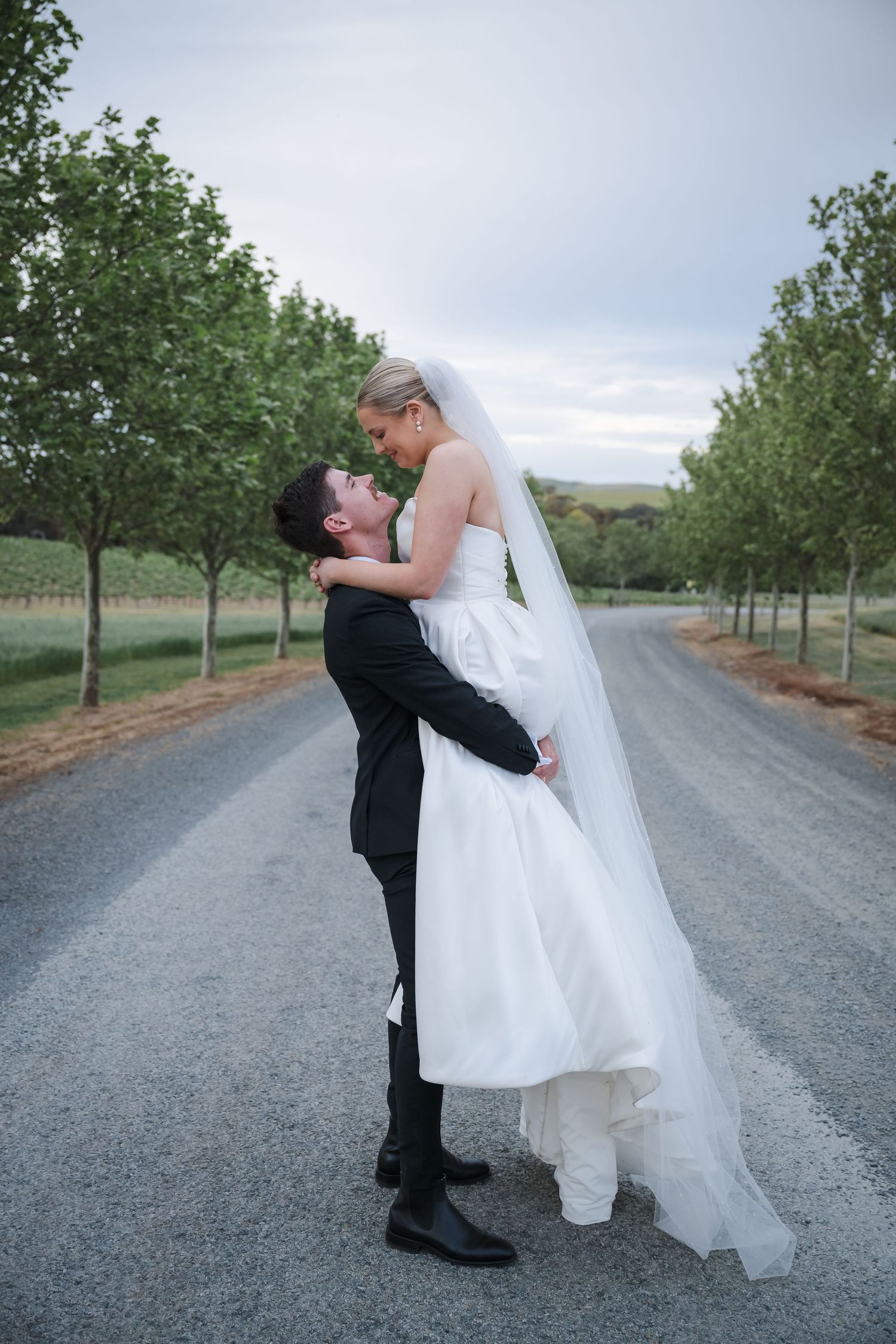 Romantic wedding couple embraces on a tree-lined gravel road during their countryside photo session.