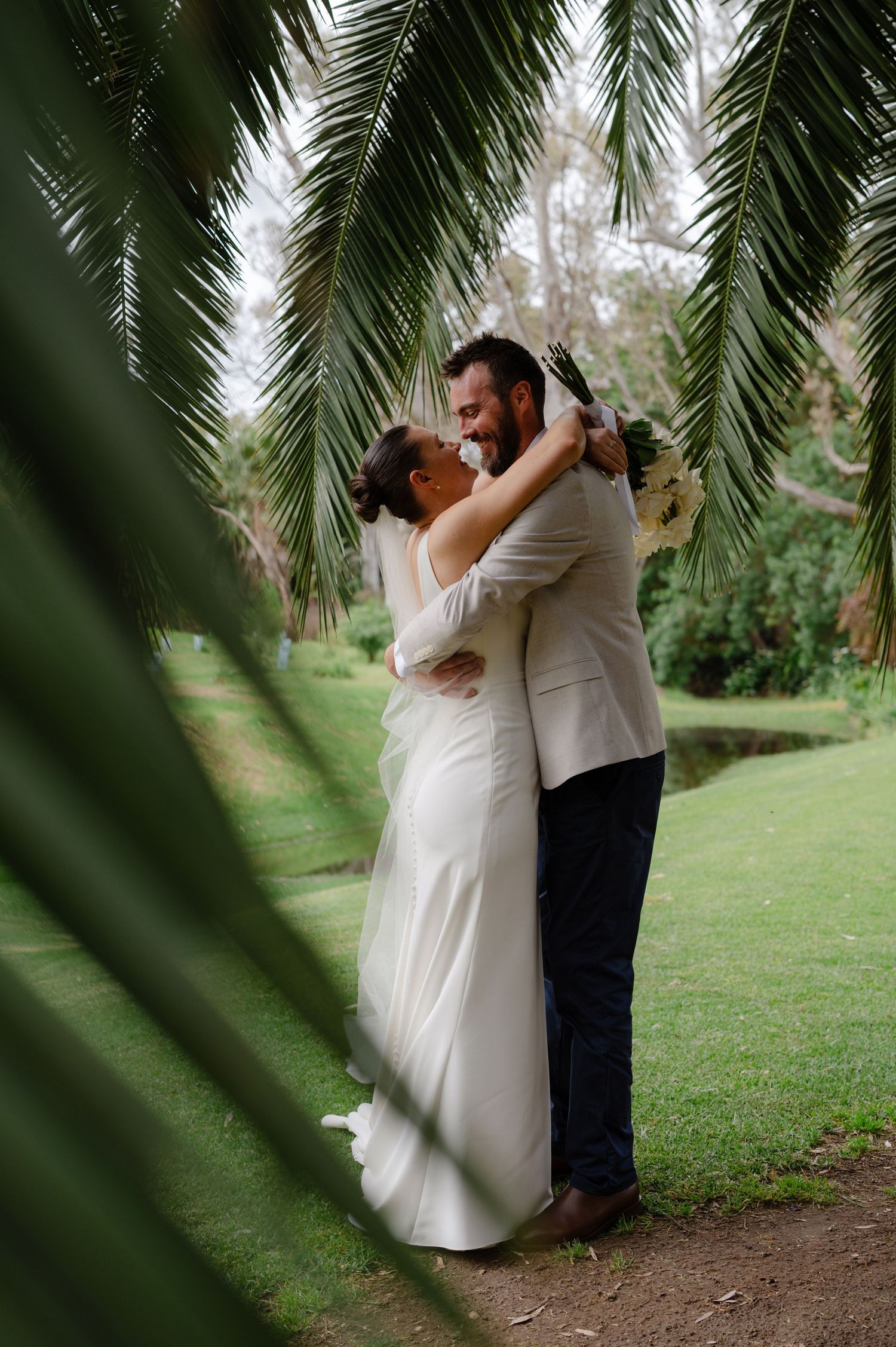 A romantic wedding couple embraces under tropical palm fronds in a lush garden setting.