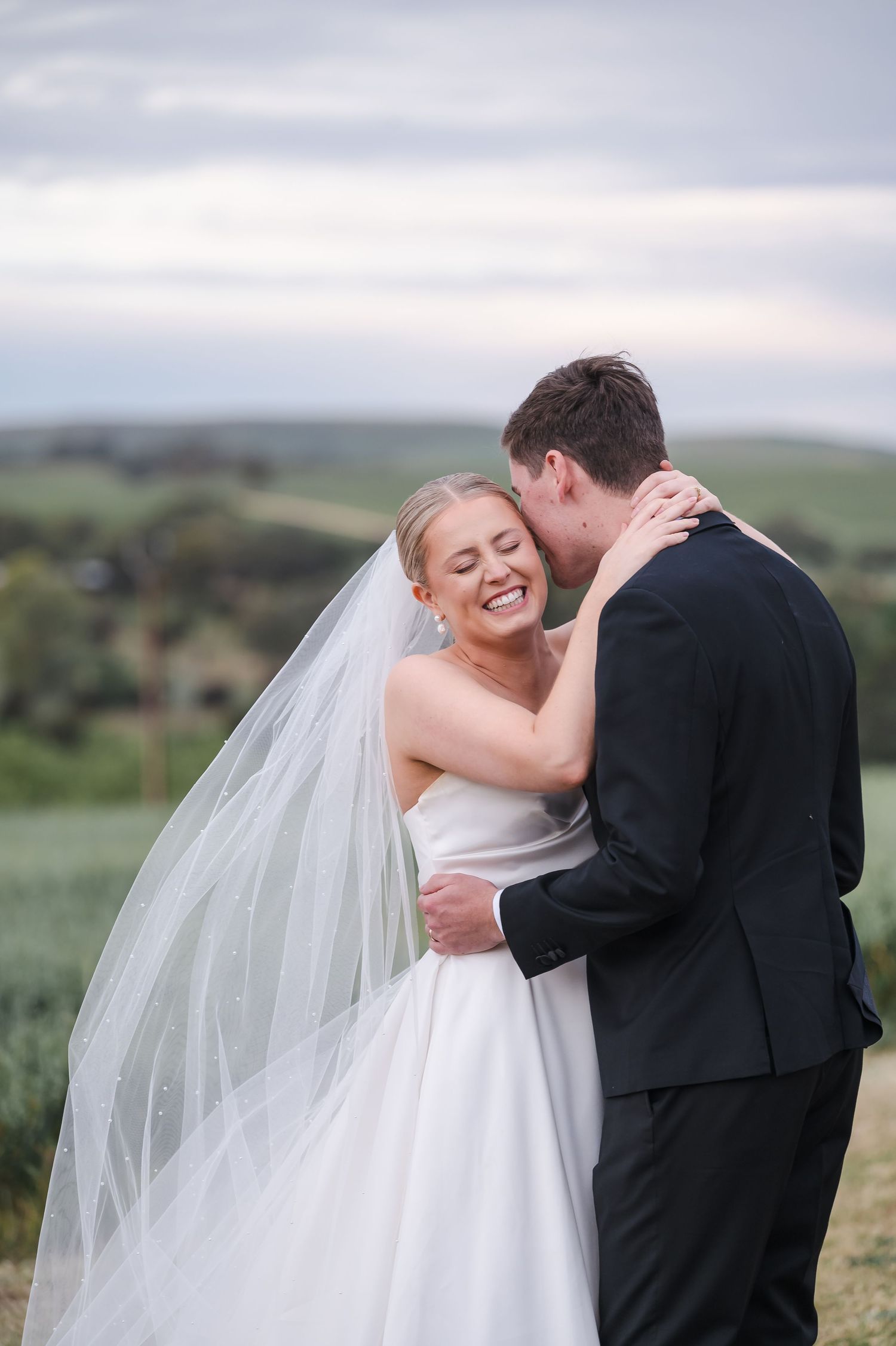 A newlywed couple embraces joyfully against a rolling countryside backdrop during their wedding photoshoot.