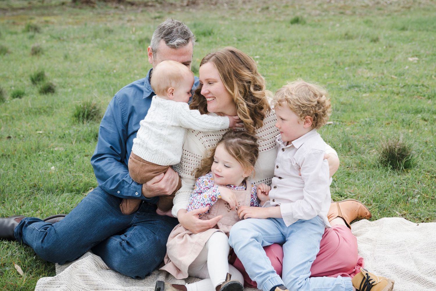 A loving family sits together on a blanket in a grassy field during an outdoor photo session.