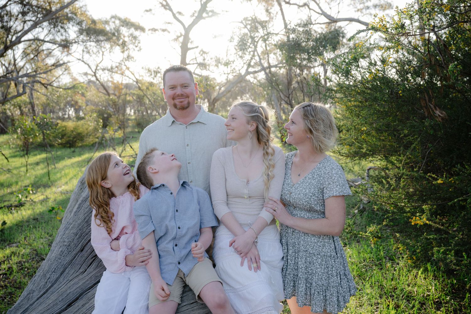 A family group sits together on grass in a sunlit outdoor setting with trees and greenery in the background.
