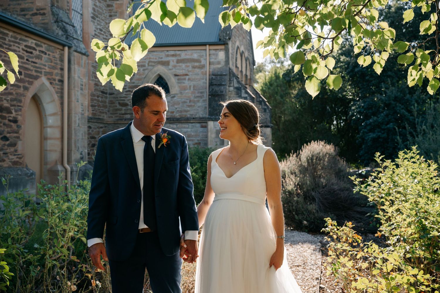 Couple in wedding attire walks together on a sunny day through a stone courtyard with green foliage overhead.