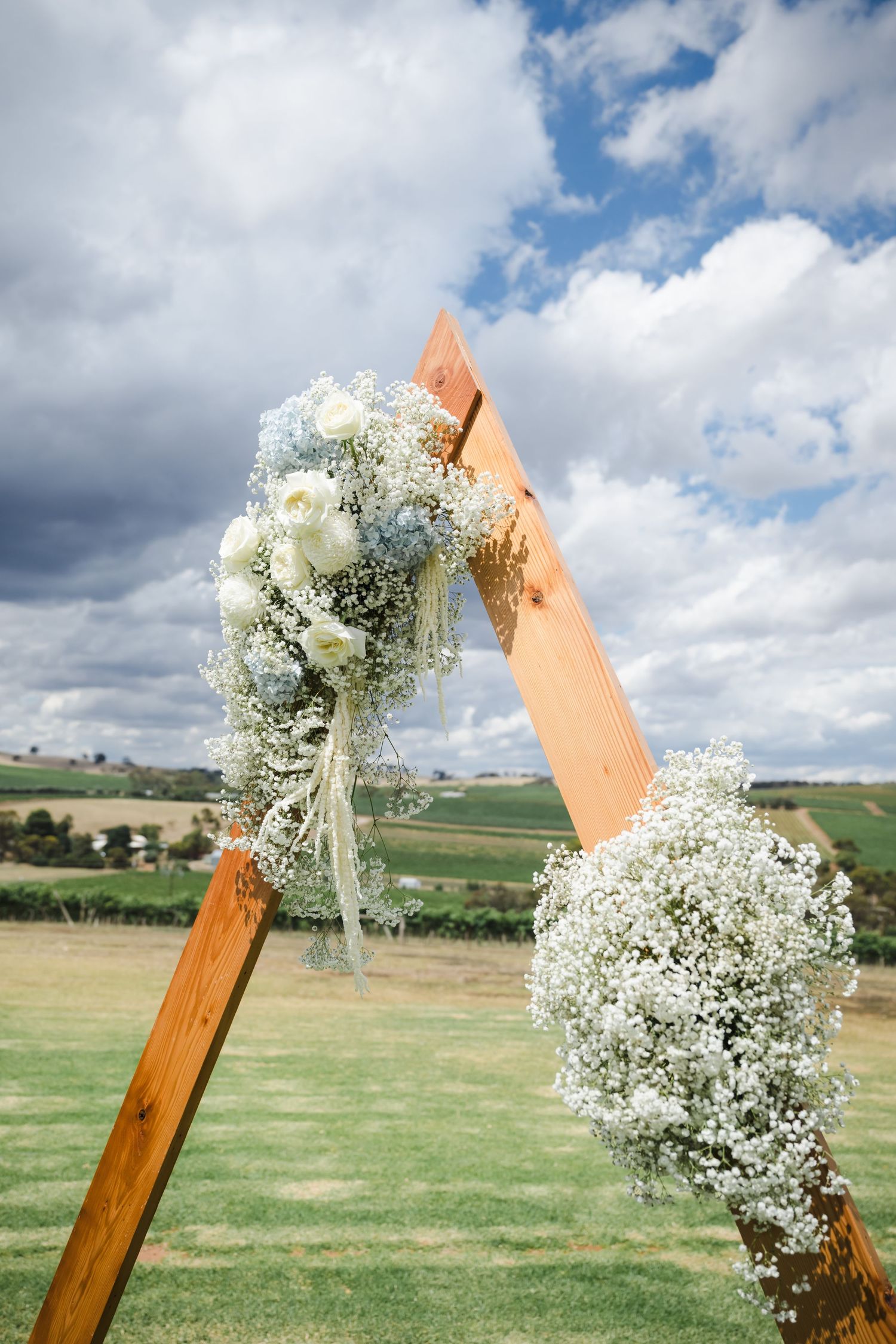 Wooden triangular wedding arch decorated with white baby's breath flowers and roses against a cloudy O'Leary Walker