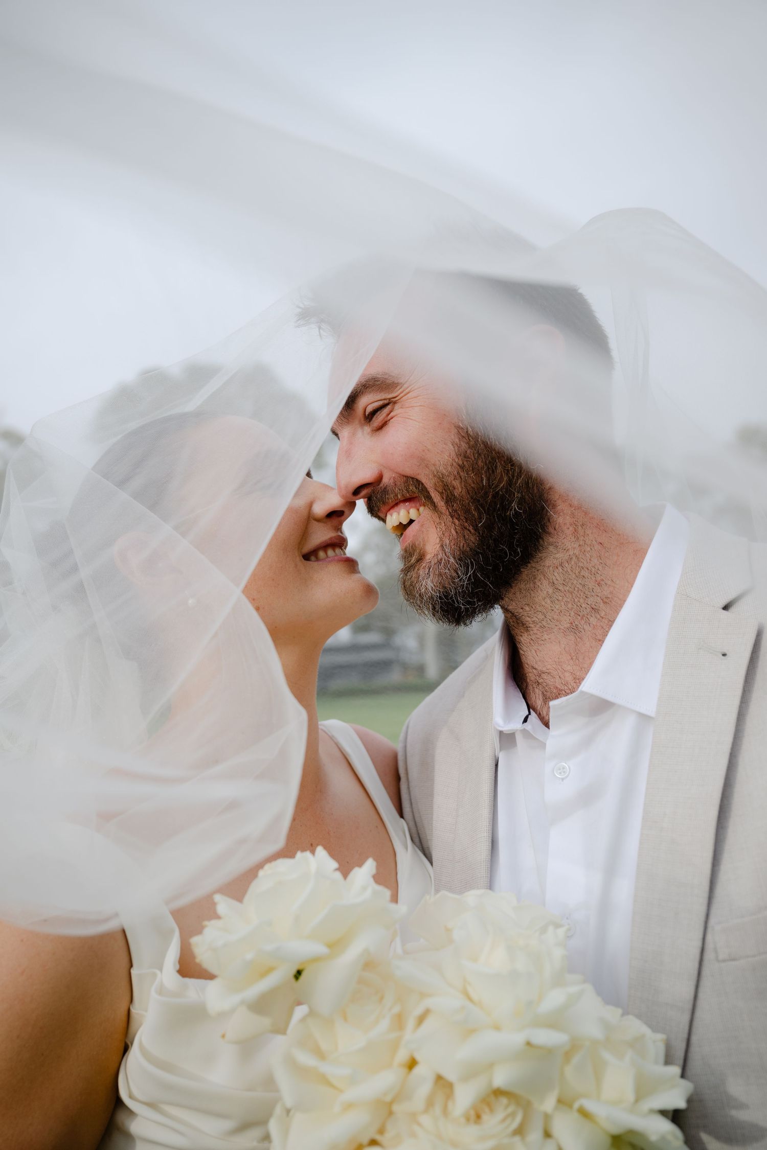 A romantic wedding moment captured under a flowing white veil with cream roses in the foreground.