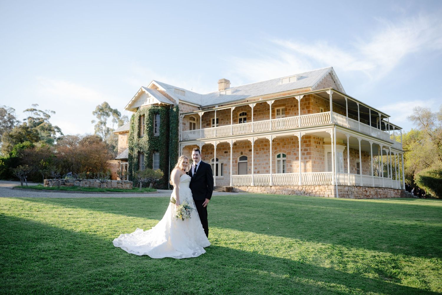 A couple stands together on a lush green lawn in front of a grand historic mansion with white wraparound porches.
