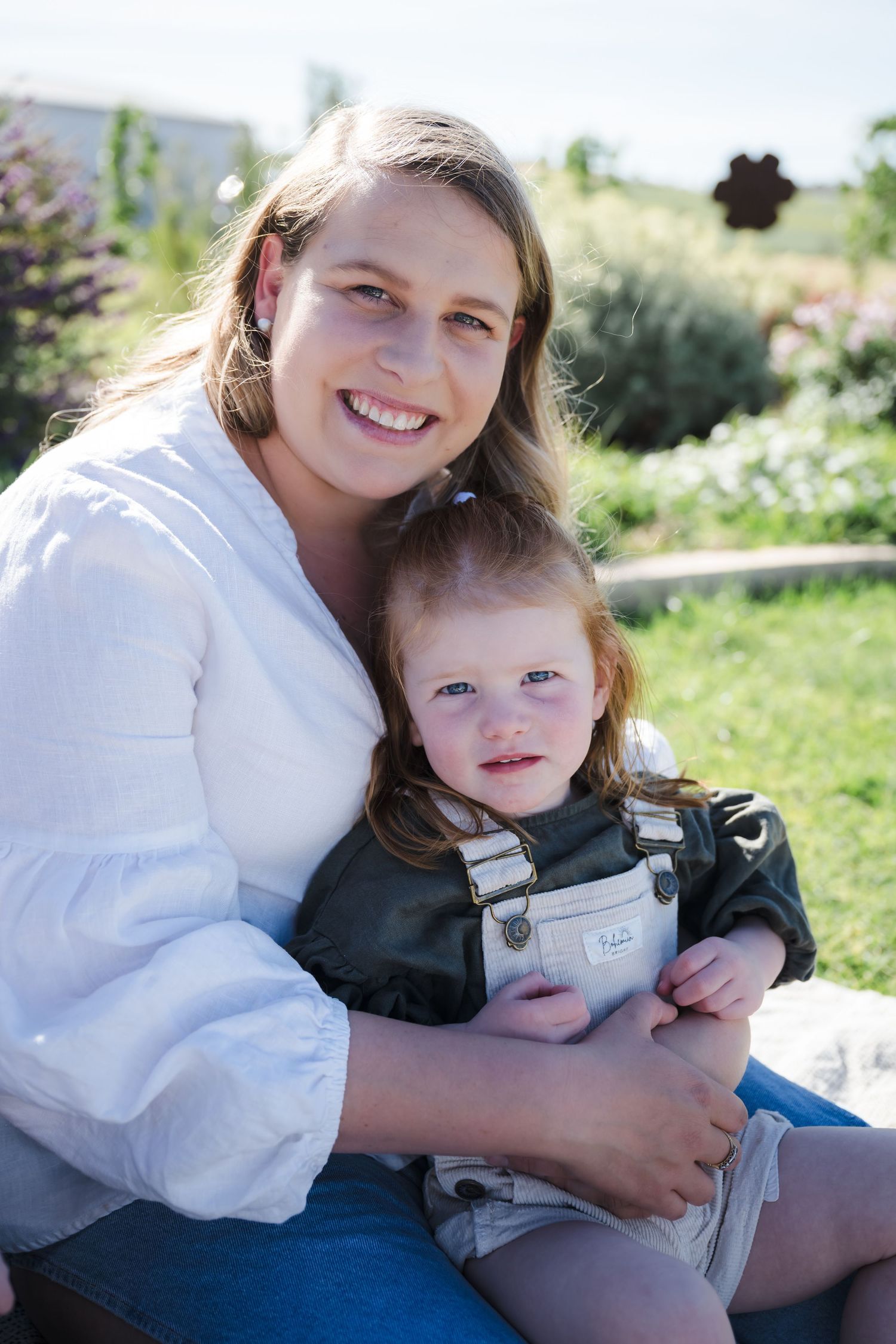A parent and small child sit together outdoors on a sunny day in a garden setting.