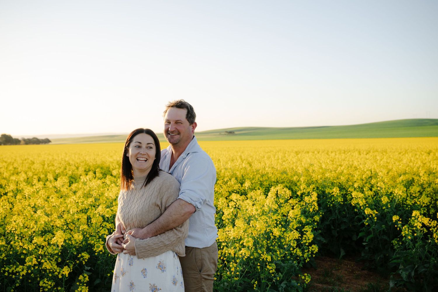 Happy couple embracing in a bright yellow flowering canola field at sunset.