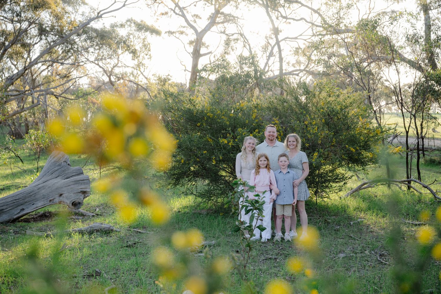 A family in white and blue outfits poses together in a sunlit meadow with yellow wildflowers and trees.