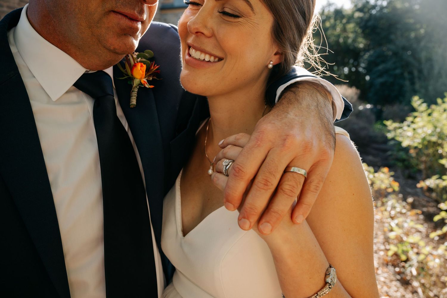 Close-up shot of newlyweds showing wedding rings and boutonniere in warm sunlight.