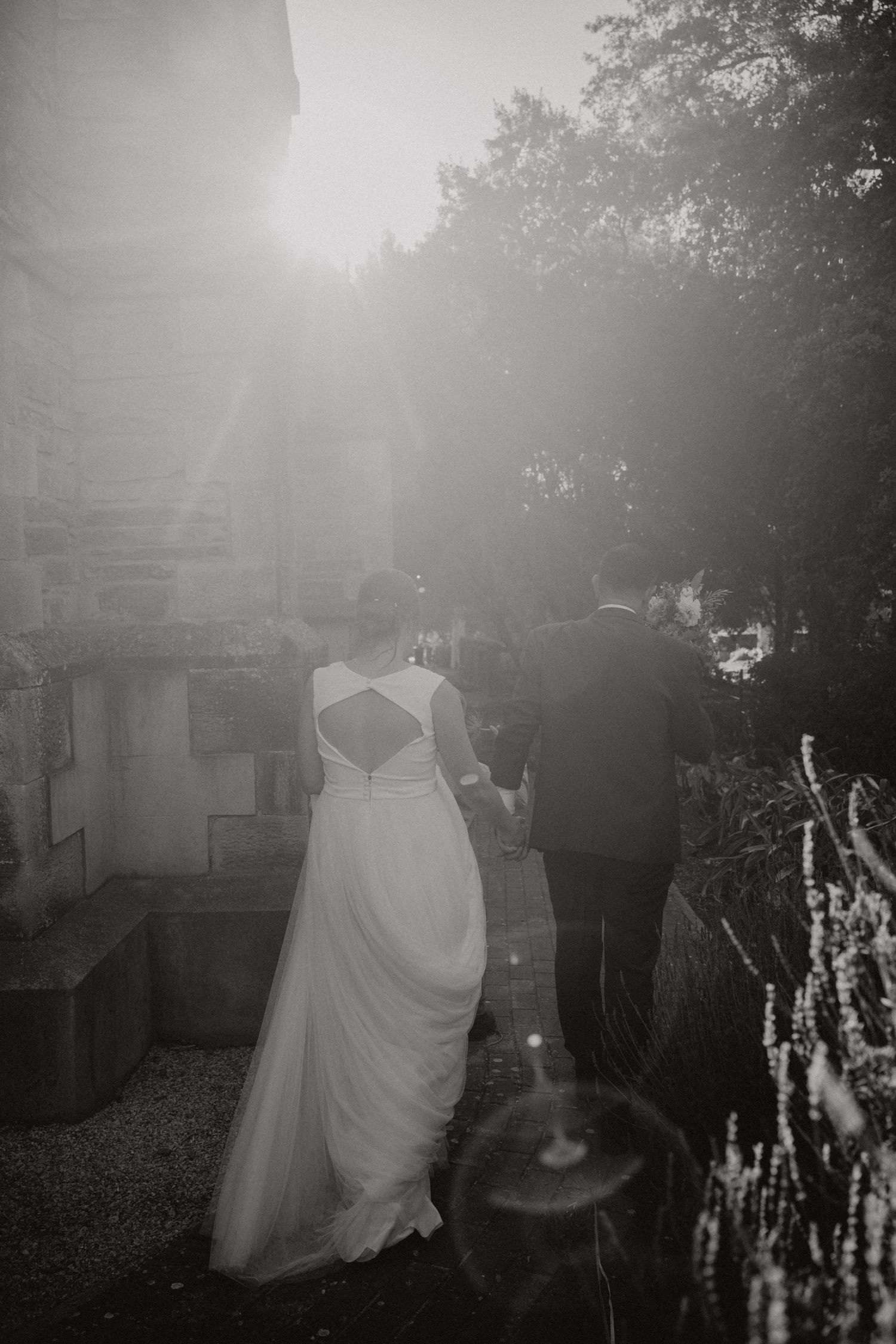 A dreamy black and white wedding portrait with sunlight streaming through trees creating ethereal atmosphere.