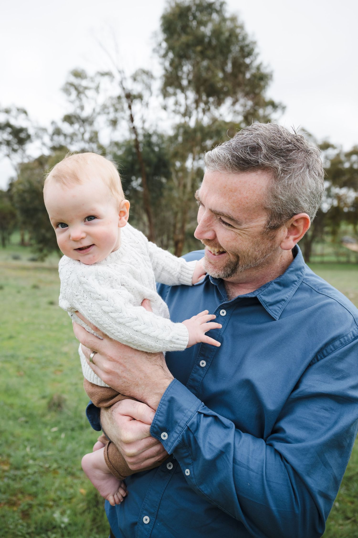 Parent in blue shirt holds and plays with baby wearing white sweater in outdoor setting with trees in background.