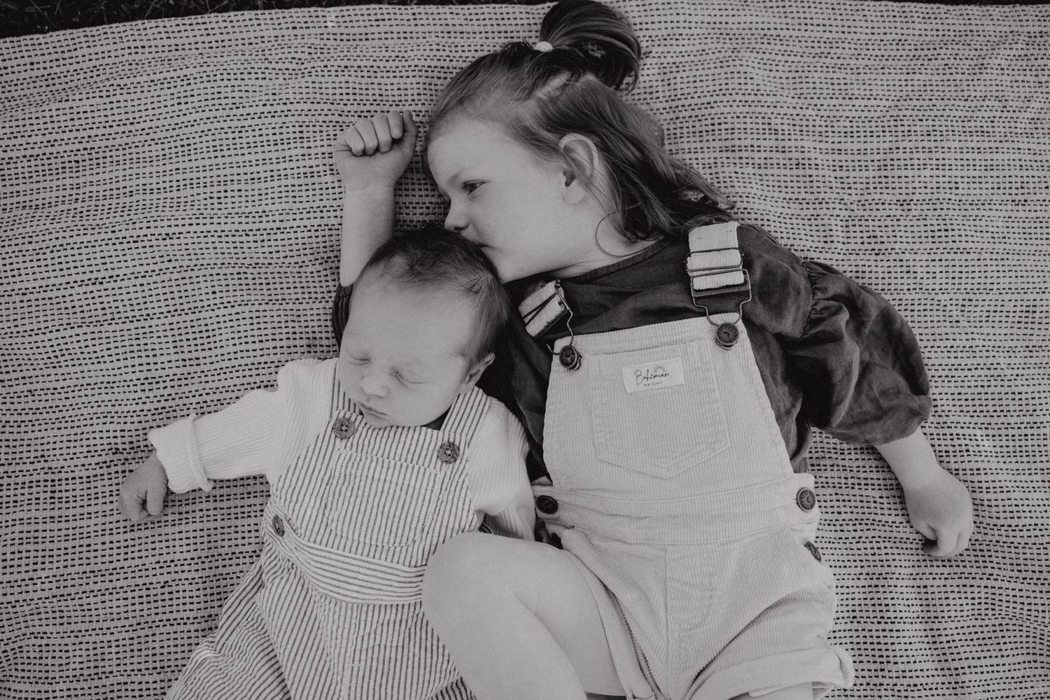 A black and white photo shows two people wearing matching overalls lying together on a textured surface.