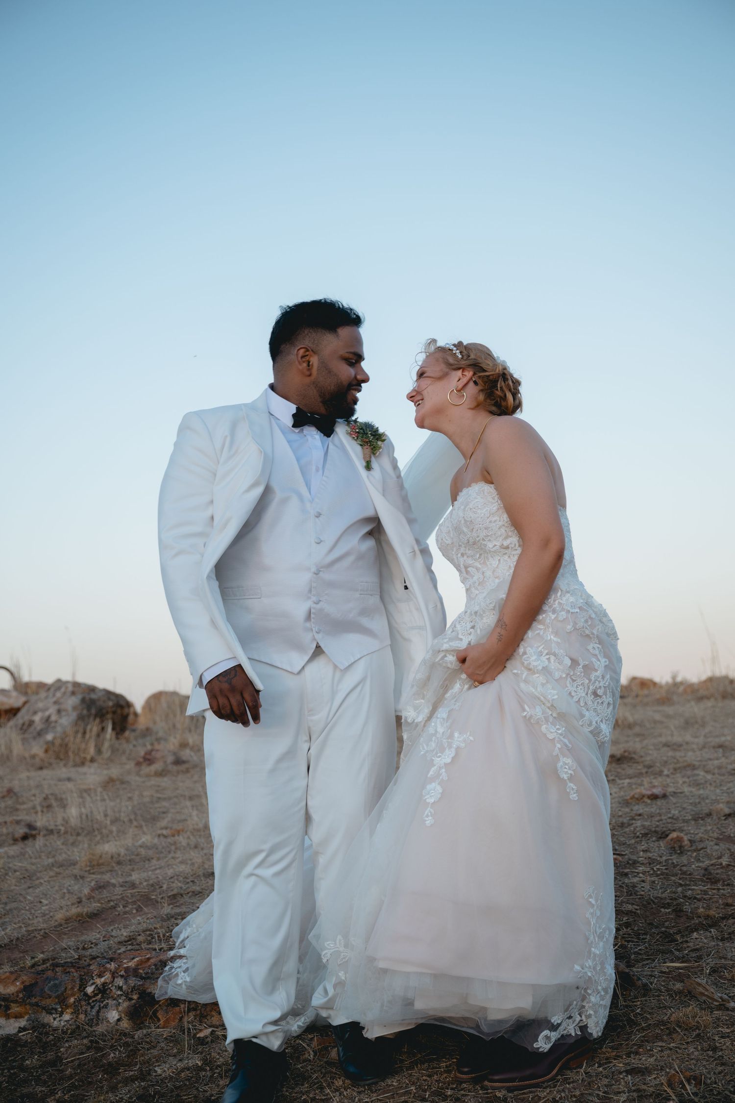 A couple in matching white wedding attire share intimate moments during a sunset photoshoot in an outdoor setting.