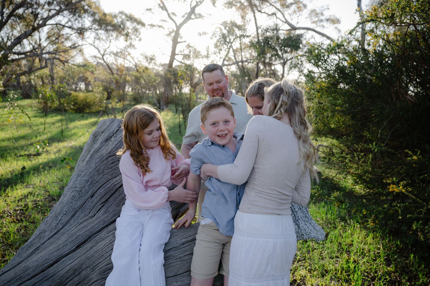 A joyful outdoor family moment captured on a blanket in a sunlit grassy field with trees in the background.