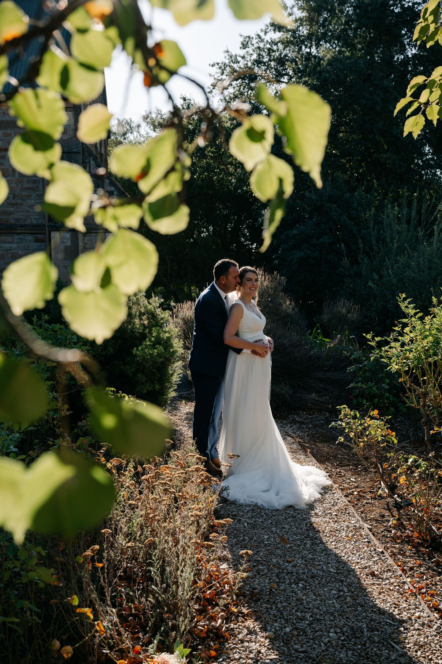 A couple in wedding attire embrace on a sunlit garden path framed by golden autumn leaves.