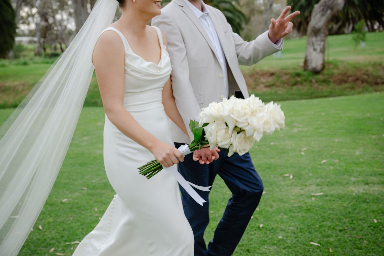 A couple in wedding attire walks on grass, with the bride holding a white bouquet and wearing a flowing veil.
