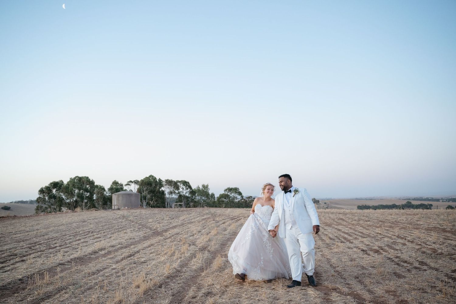 A couple in white outfits walk hand in hand along a serene beach during sunset.