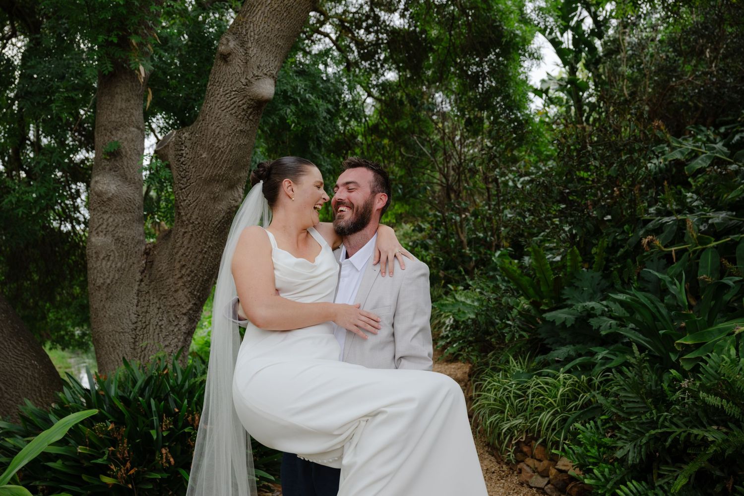 A couple in wedding attire shares an intimate moment surrounded by lush greenery in a garden setting.