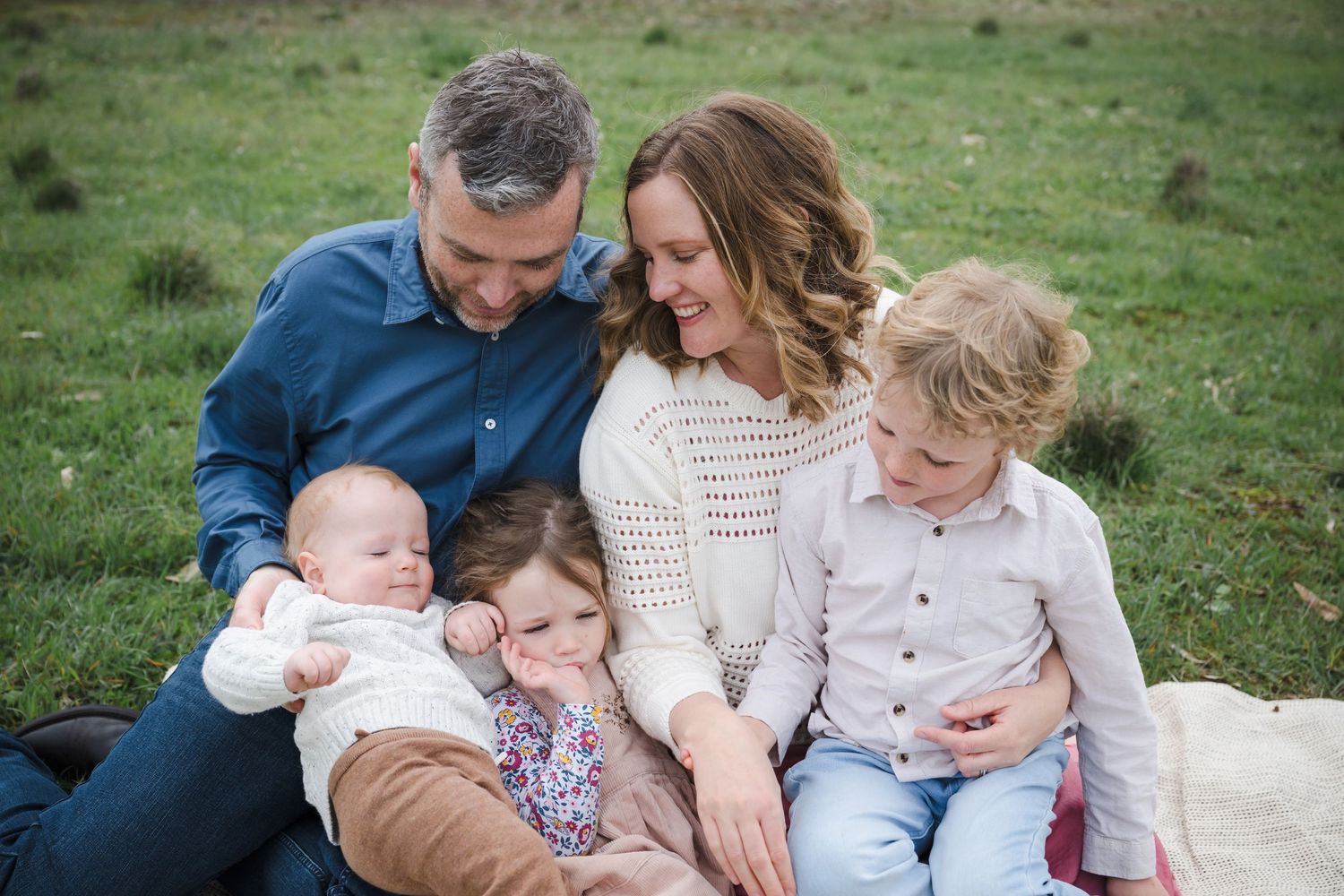 A family sits together on a blanket in a green field, sharing a tender moment outdoors.