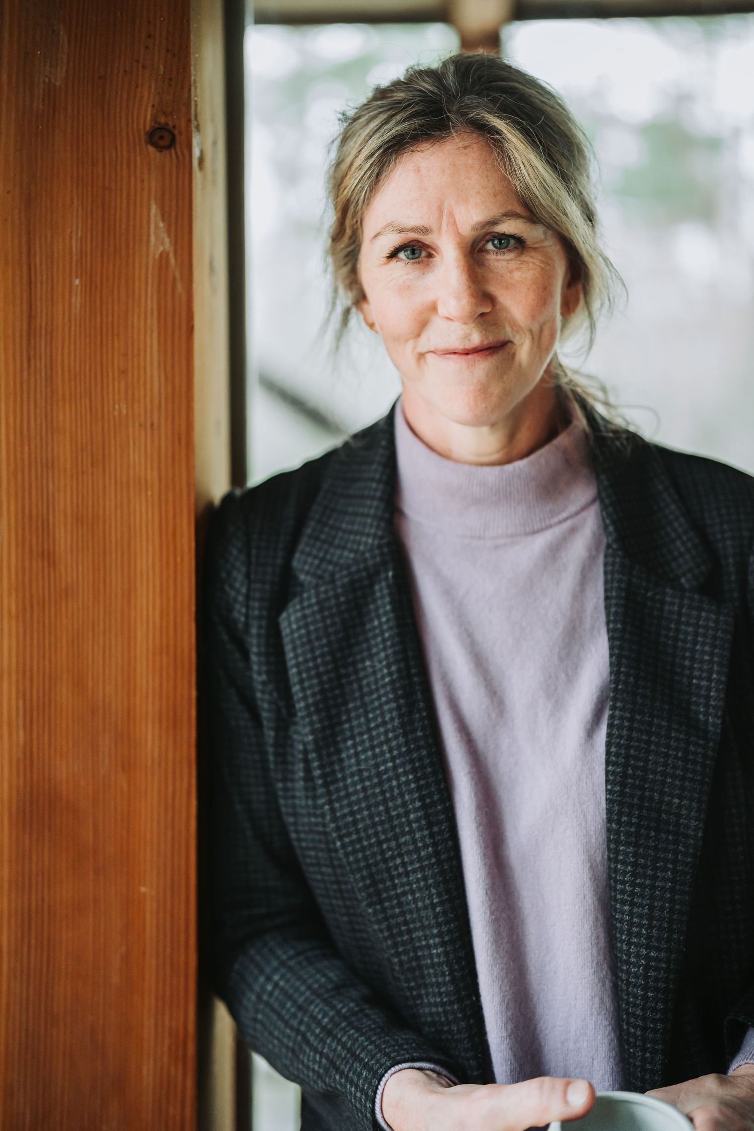 Person wearing a black blazer and light turtleneck sweater poses against wooden window frames in a portrait series.