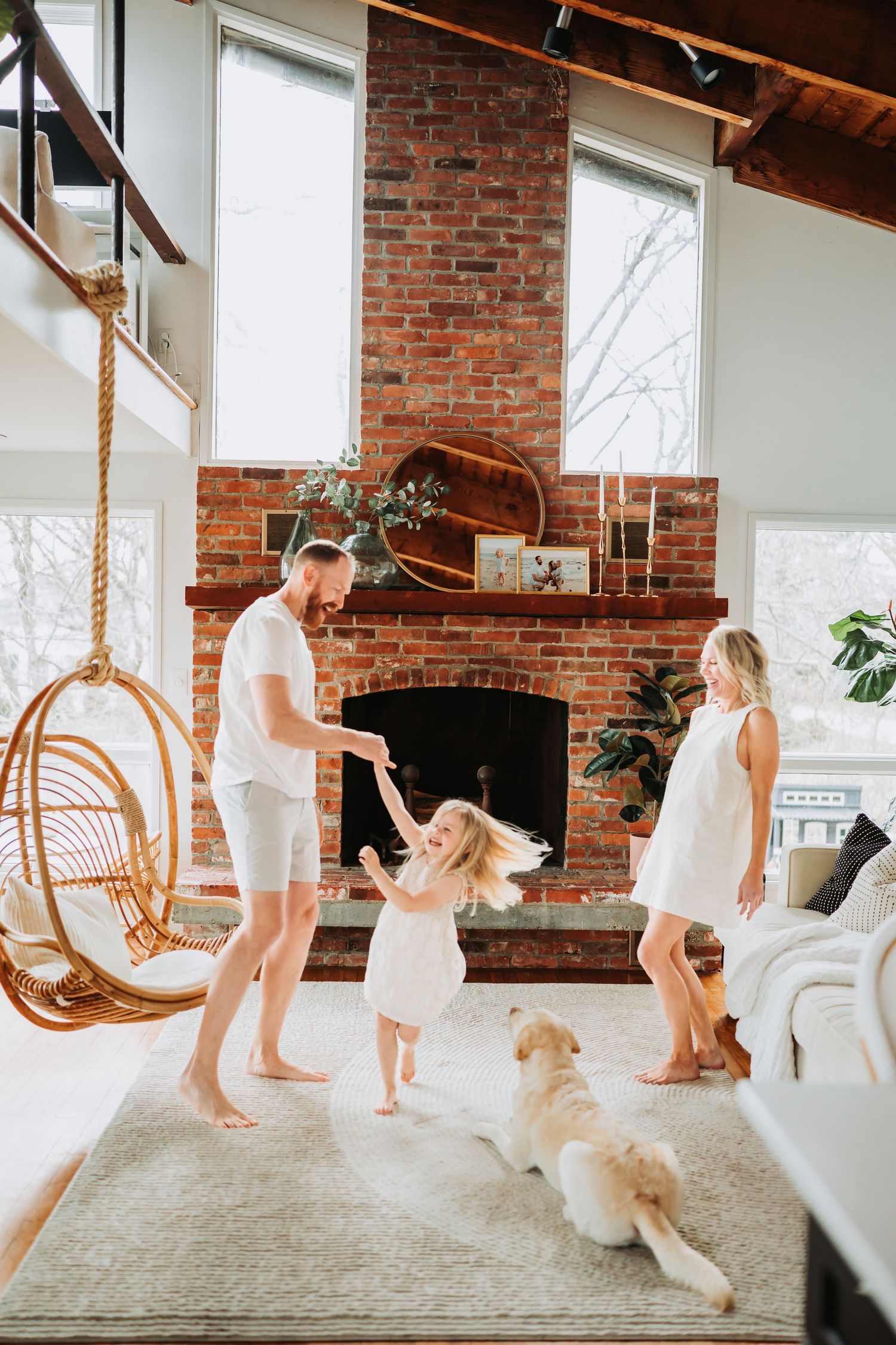 Family dancing in bright living room with brick fireplace, hanging rattan chair, golden retriever, and large windows.