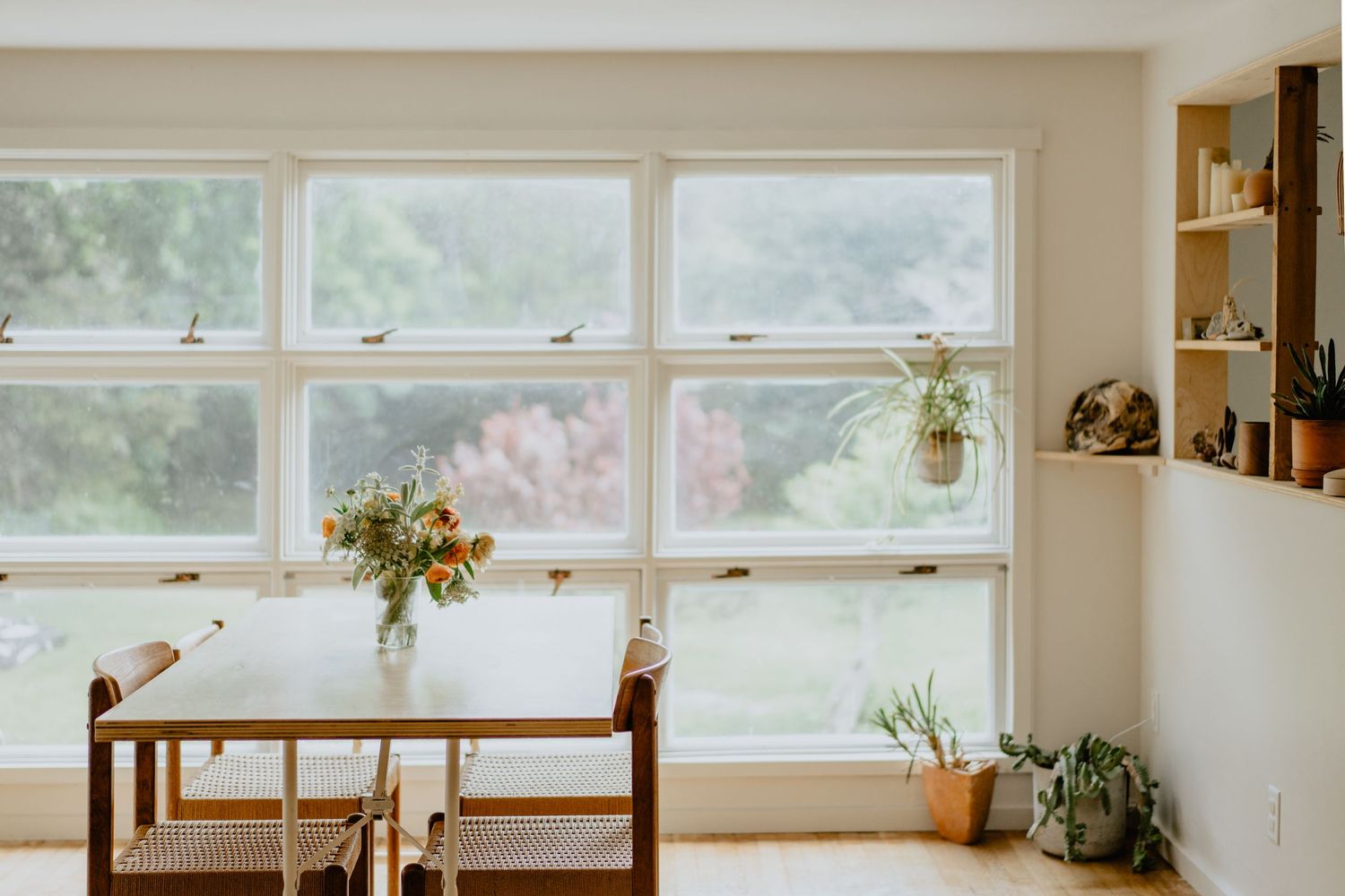 Bright dining area with large windows, wooden table, and hanging plants in natural light.