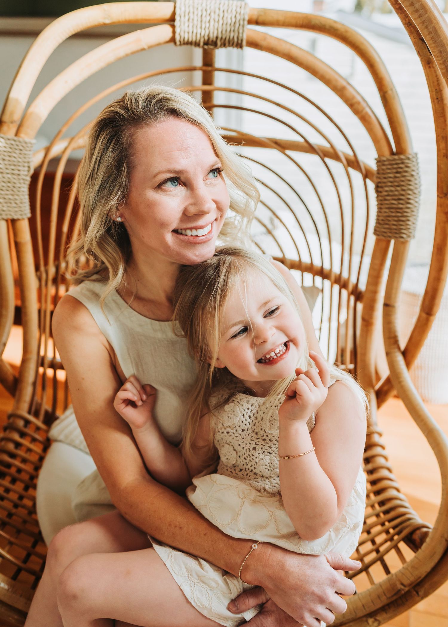 Smiling mother and toddler daughter sitting together in a wicker rattan chair wearing white dresses.