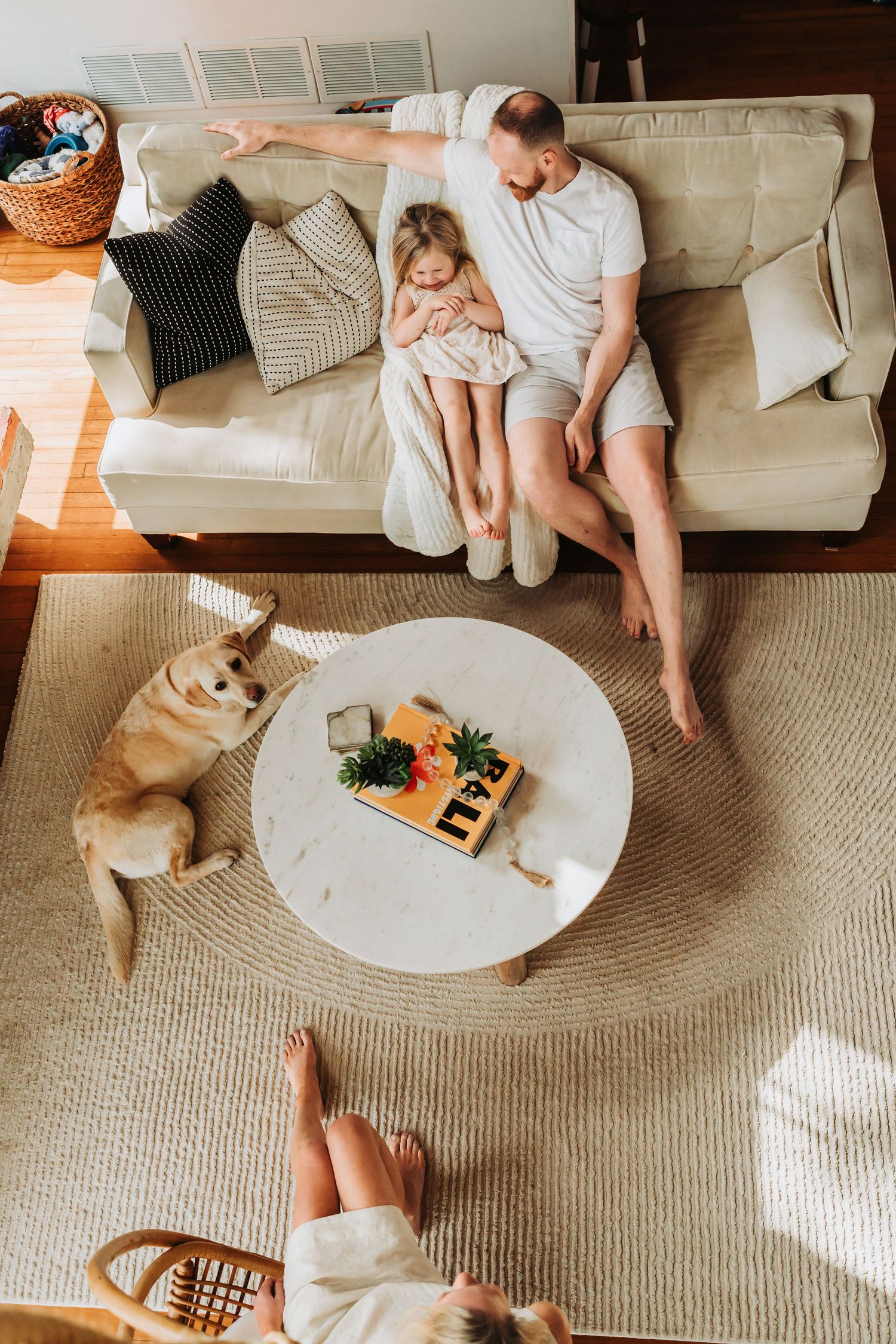 Father cuddling daughter on beige sofa with golden retriever nearby on woven rug in sunny living room.