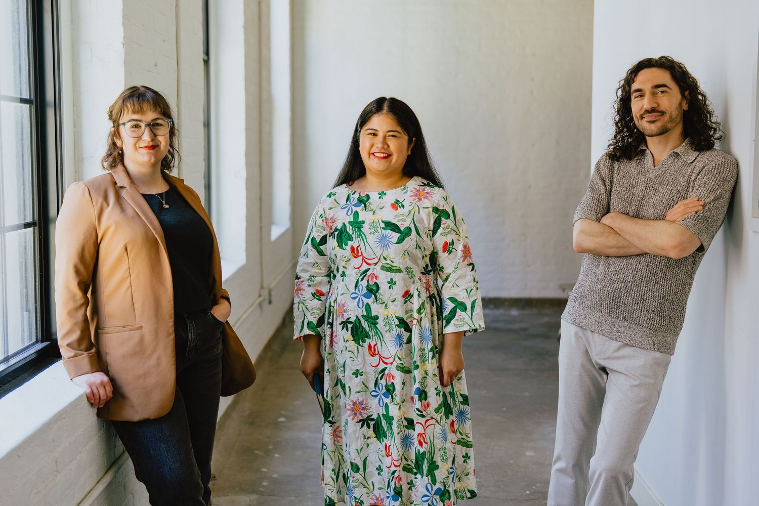 Three colleagues stand casually by large windows in an office hallway.