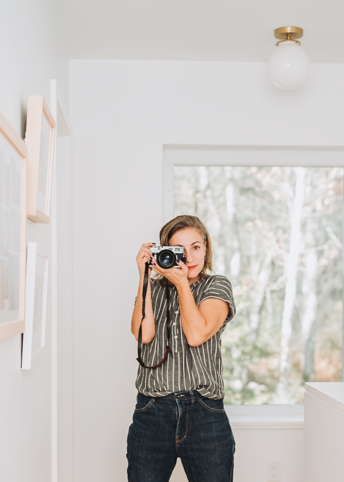A photographer in a striped shirt and dark jeans takes a picture with a vintage camera in a bright white room.