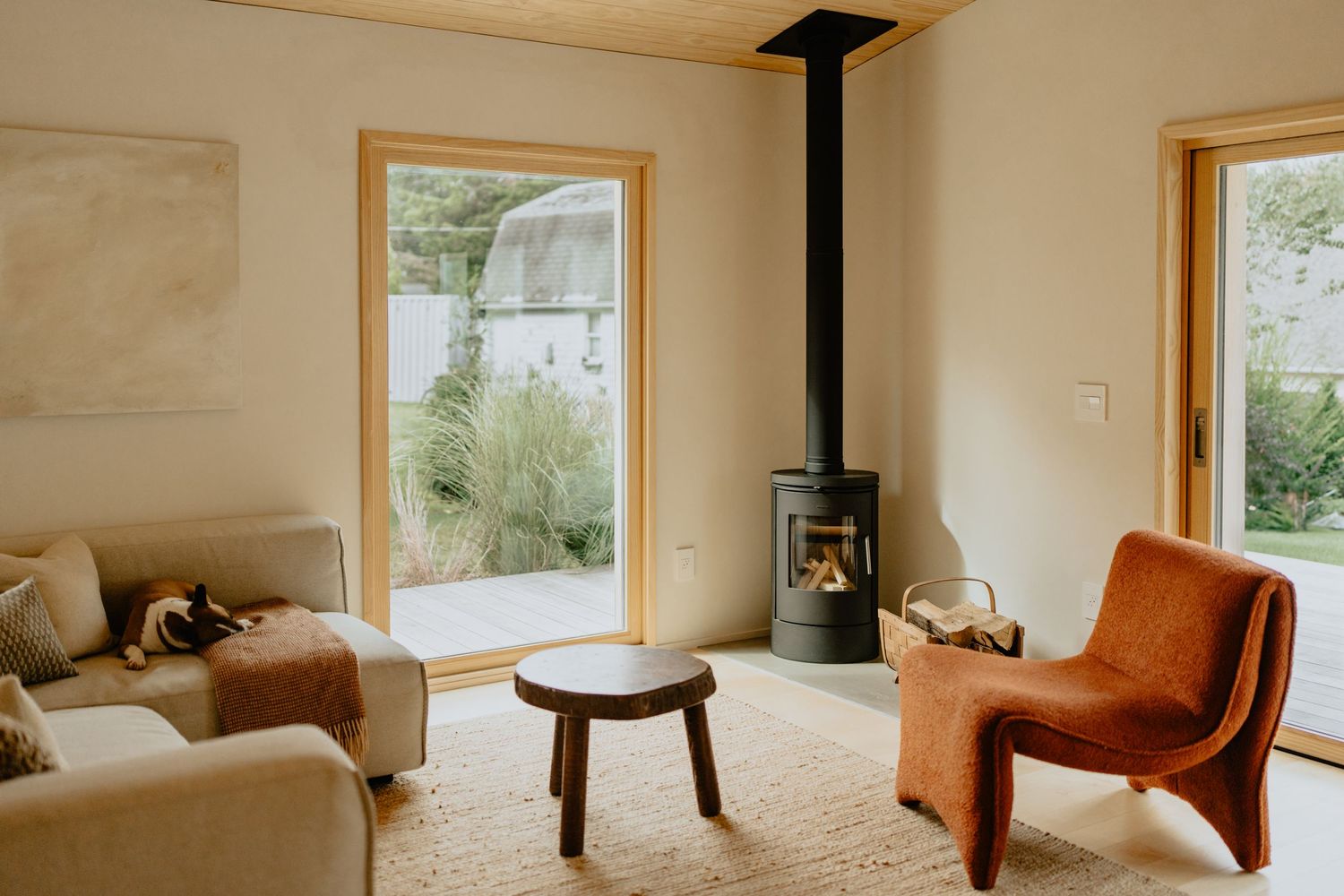 Modern minimalist living room with wood stove, orange chair, and large windows overlooking nature.