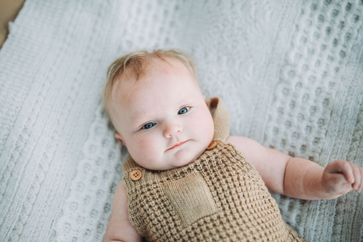 A baby wearing a knitted tan sweater lies on a textured gray blanket while looking up at the camera.