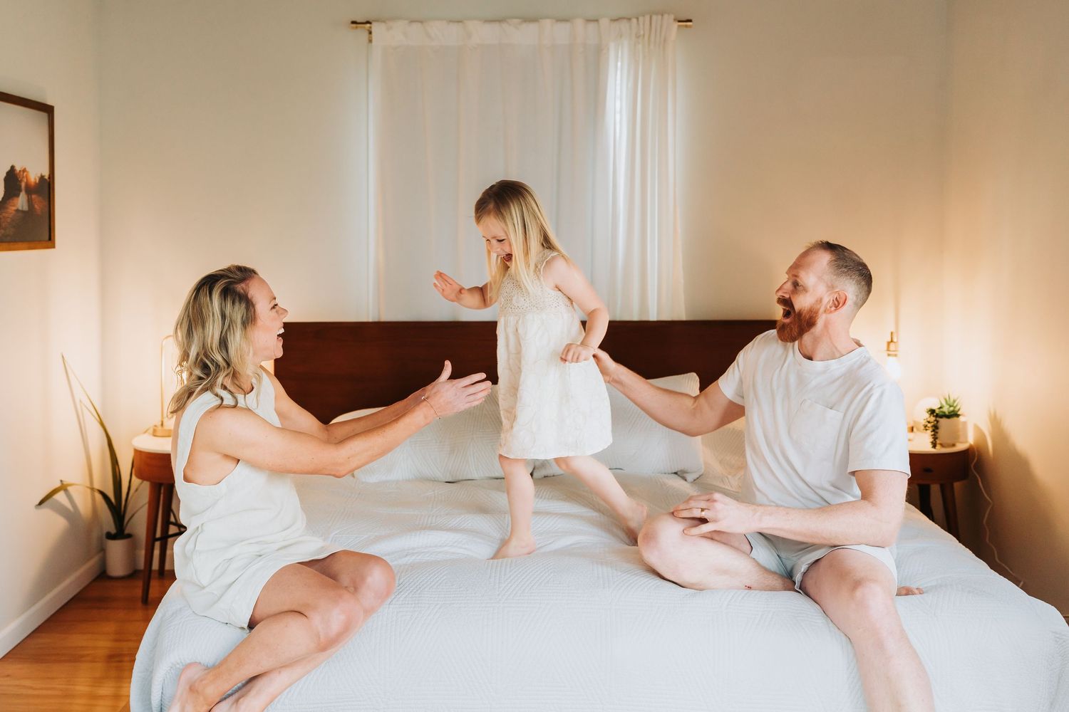 A happy family plays on a white bed, with a toddler girl standing between her smiling parents in a bright bedroom.
