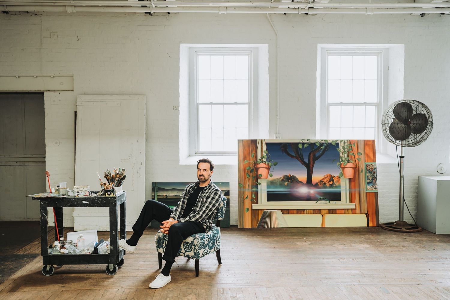Artist sits in a chair in a minimalist studio space with white walls, windows, and artwork displayed against the wall.