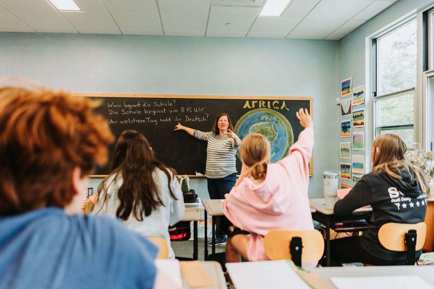 Students raise their hands to participate during a geography lesson with Africa displayed on the chalkboard in a classroom.