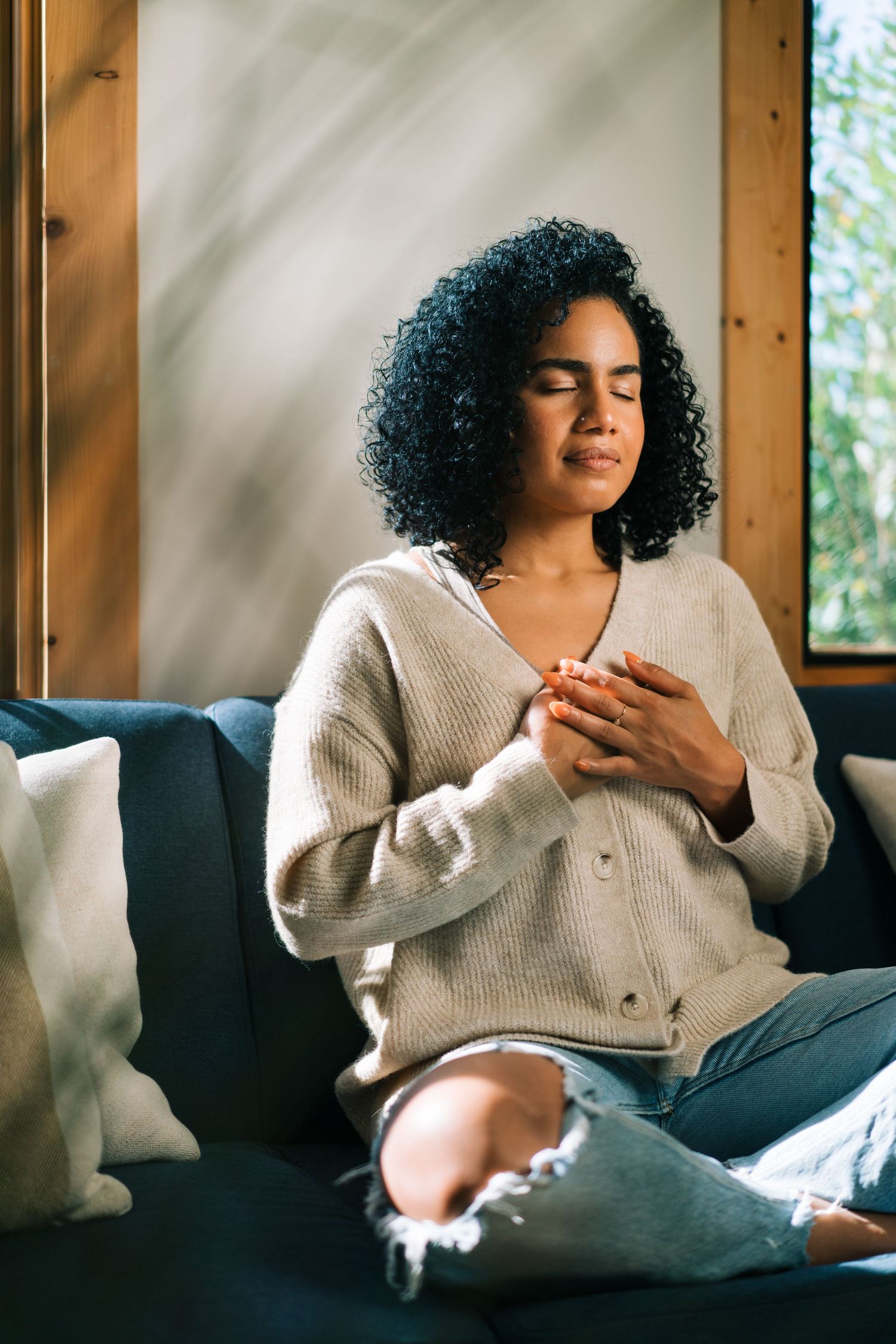 Woman in beige cardigan sitting on couch with eyes closed in peaceful moment.