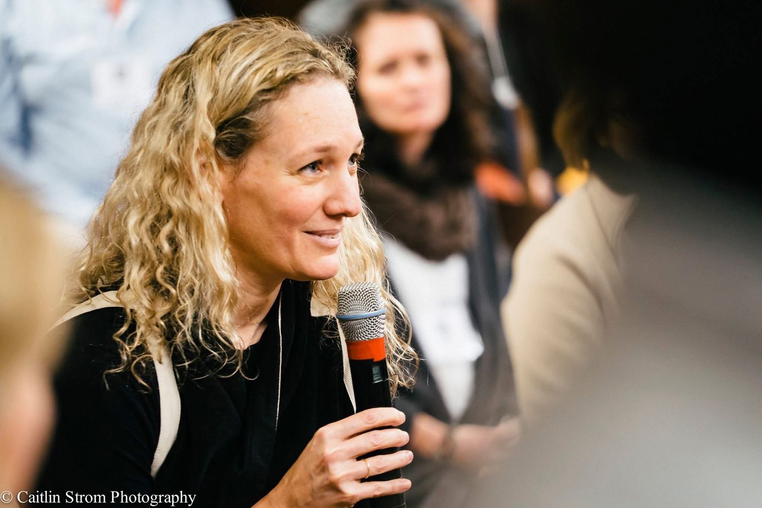 A person with blonde curly hair drinks from a paper coffee cup while sitting outdoors.