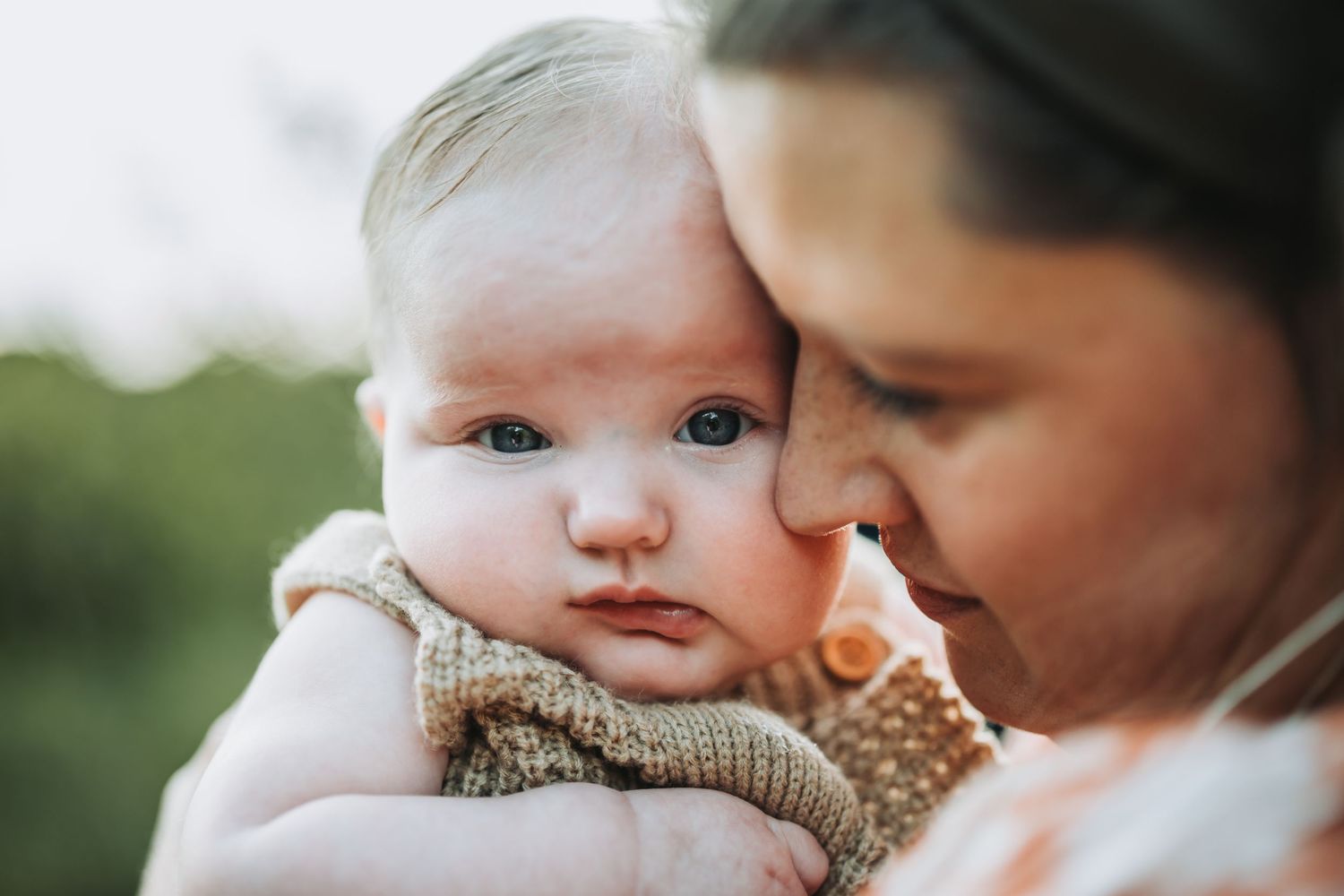 A tender moment shows a parent and baby sharing a gentle kiss outdoors in natural lighting.
